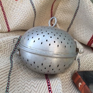 May include: A spherical, silver-toned metal tea infuser featuring a perforated surface, a hinged closure mechanism, and a small top loop for handling. The metal body displays faint embossed text near the center seam, indicating its functional design.