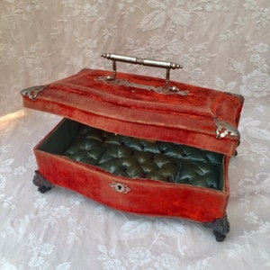 May include: A rectangular jewellery box covered in red velvet with a hinged lid, featuring a silver-toned metal handle, decorative corner accents, and four ornate feet. The interior is lined with tufted green satin and includes a keyhole plate.