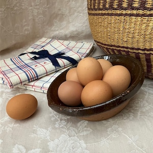 May include: A dark brown ceramic bowl filled with several light brown eggs, with one additional egg resting nearby. Beside the bowl is a folded white cloth napkin featuring a blue and red plaid pattern, tied with a dark blue ribbon.
