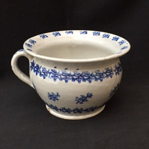 May include: A white ceramic chamber pot with a handle and blue floral patterns. The rim and body are decorated with blue floral patterns. The pot has a round base and a wide opening.