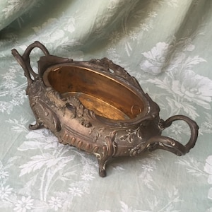 Puede incluir: Una fuente antigua de metal color bronce con un interior ovalado y detalles florales ornamentados. La fuente tiene dos asas curvas y descansa sobre cuatro patas pequeñas. El interior tiene un acabado dorado.