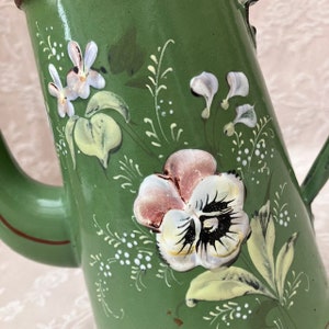 May include: A green enameled metal pitcher featuring hand-painted floral motifs. The design includes a large central pansy with white, pink, and dark purple petals, surrounded by smaller white blossoms, green leaves, and delicate white dotted accents.