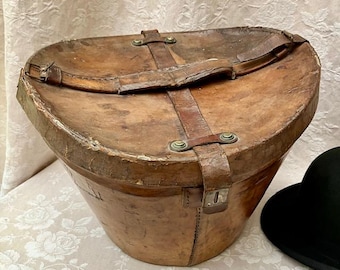 May include: A weathered brown leather hat box with a rounded triangular shape, featuring a central leather strap, metal rivets, and a buckle closure. The exterior shows signs of age and wear, with a sturdy handle attached to the top lid.