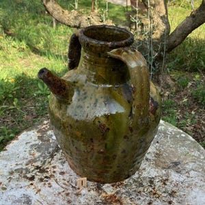 Puede incluir: Una jarra de cerámica antigua, vidriada en verde, con pico y asa. La jarra tiene un cuerpo bulboso y una abertura ancha. El esmalte tiene un aspecto moteado con variaciones de color. Es probable que sea una pieza de cerámica antigua.