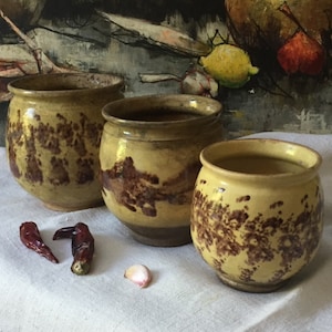 Può includere: Tre vasi in ceramica rotondi con smalto giallo e macchie marroni. I vasi variano in dimensioni e sono disposti in gruppo. Due peperoncini rossi essiccati e uno spicchio d'aglio sono posizionati sulla superficie in tessuto bianco.