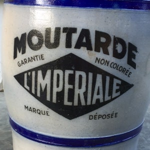 May include: A vintage, off-white ceramic mustard jar with a blue rim and accents. The jar features the text "MOUTARDE L'IMPERIALE" in black, with additional text indicating a guarantee and trademark.