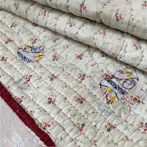 Puede incluir: Un textil acolchado con una base de color blanco roto y un delicado estampado floral en rojo, amarillo y verde. La tela está cosida con hilo rojo en un patrón de cuadrícula e incluye dos parches distintos con motivos florales abstractos.