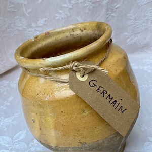 Puede incluir: Una vasija de cerámica esmaltada en amarillo con un borde ancho y cuerpo redondeado. Un trozo de cuerda marrón está atado alrededor del cuello de la vasija, sosteniendo una etiqueta de papel marrón con la palabra GERMAIN escrita en tinta negra.