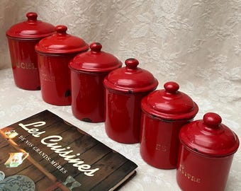 May include: A set of six red enamel kitchen canisters with lids, arranged in a row. The canisters have labels in French and are accompanied by a vintage cookbook titled "Les Cuisines de nos Grands-Mères."