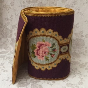 May include: A rolled-up, decorative needlepoint fabric in shades of burgundy, gold, and light blue. The fabric features a central floral design with a pink rose and green leaves, framed by an oval border.