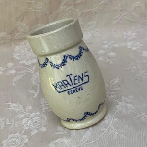 May include: An off-white ceramic jar with a slightly tilted design. The jar features blue floral garland detailing and the text "MARTENS GENEVE" in blue. The jar has a wide opening and a small base.