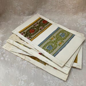 May include: A stack of fabric samples featuring embroidered floral patterns. The samples are attached to white cardstock with staples. The embroidery includes red, yellow, green, and blue colors. The background is a white floral patterned fabric.