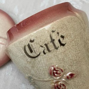 May include: Cream-colored ceramic coffee cup with the word "Cafe" in black script. The rim is a faded red, and the cup features embossed pink roses. The cup has a crackle glaze.