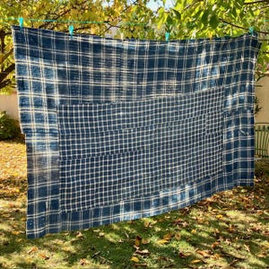 Può includere: Un grande tessuto a quadri blu e bianco appeso a un filo per stendere i panni. Il tessuto ha un design patchwork con un motivo a griglia ripetuto. Il tessuto è sospeso con mollette verdi, con un prato erboso e fogliame sullo sfondo.