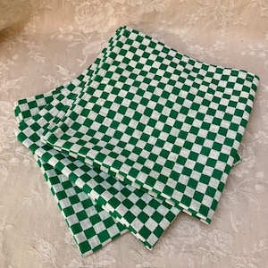 May include: A set of three square cloth napkins featuring a repeating green and white checkered pattern. The napkins are folded and stacked, displaying a consistent grid design across the fabric surface with visible stitched edges on all sides.