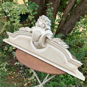 May include: A light gray architectural pediment featuring a central shield emblem, scrollwork, and radiating fan-like carvings. The elongated, horizontal decorative element has a smooth finish and symmetrical molded edges along the top and base.