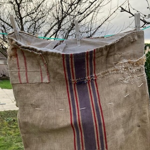 May include: A vintage beige linen bag with red and purple stripes, hanging on a clothesline with white clothespins. The bag has a rectangular patch on the side and shows signs of wear and tear, adding to its rustic charm.