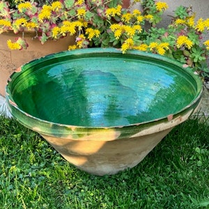 May include: A large, antique green glazed ceramic planter with a wide, shallow bowl and a conical base. The planter has a distressed, aged appearance, with visible wear and tear. The background features green grass and yellow flowers.