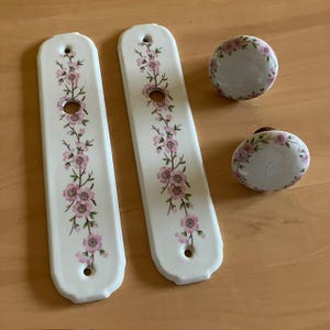 Peut inclure: Deux plaques de porte rectangulaires en céramique blanche et deux boutons de porte ronds assortis, ornés d'un motif floral rose délicat avec des tiges et des feuilles vertes. Chaque plaque présente une ouverture centrale et deux trous.