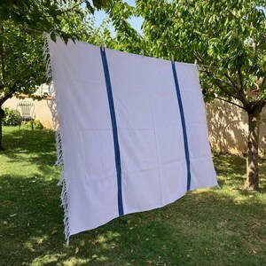 May include: White rectangular textile with blue vertical stripes and fringed edges, hanging on a clothesline outdoors. The textile is suspended between trees in a grassy area, suggesting a decorative or functional item.