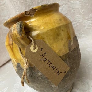Pode incluir: Um pote de cerâmica rústico com a parte superior esmaltada em amarelo e a base cinzenta texturizada. A borda apresenta uma lasca visível. Uma etiqueta de papel castanho atada com cordel à asa lateral contém o texto "ANTONIN".