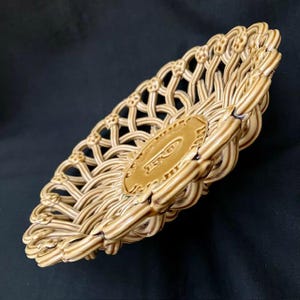 Könnte beinhalten: Beige Keramikkorb mit offenem Webmuster. Der Korb hat ein dekoratives ovales Emblem in der Mitte. Der Korb ist abgewinkelt und zeigt das komplizierte Webmuster und das geprägte Emblem.