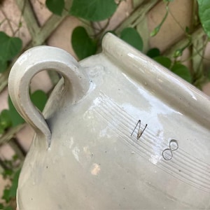 May include: A light grey glazed ceramic crock featuring a sturdy side handle and a prominent rim. The exterior surface displays the engraved characters N 8 positioned above a series of horizontal incised lines that wrap around the vessel body.