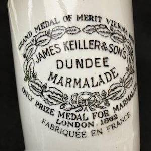 Puede incluir: Un tarro de cerámica blanca con texto impreso en negro que dice GRAND MEDAL OF MERIT VIENNA 1873, JAMES KEILLER & SON'S DUNDEE MARMALADE, ONLY PRIZE MEDAL FOR MARMALADE LONDON 1862 y FABRIQUE EN FRANCE, rodeado por una corona de hojas decorativa.