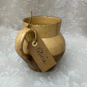 Puede incluir: Una taza de cerámica de estilo vintage, con asa, que presenta un diseño de dos tonos en tonos amarillos y marrones. Una etiqueta marrón con el nombre "Félicie" está sujeta con hilo. La taza tiene un aspecto rústico y envejecido.