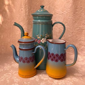 Puede incluir: Tres artículos de cocina vintage de esmalte: una cafetera alta turquesa con detalles florales, una cafetera azul y amarilla más pequeña y una jarra azul y amarilla con un patrón decorativo. Todas las piezas tienen asas y tapas curvas.