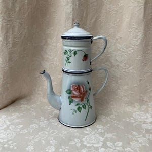 Könnte beinhalten: Eine Vintage-Kaffeekanne aus hellblauem Emaille mit floralem Design. Die Kanne hat ein zweistufiges Design mit Deckel und Griff im oberen Bereich sowie Ausguss und Griff im unteren Bereich. Das florale Design umfasst rosa Rosen und grüne Blätter.