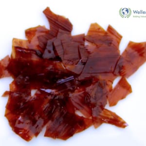 May include: A collection of irregular, translucent, dark amber-colored shellac flakes with a glossy, brittle appearance. The flakes vary in size and shape, displaying deep reddish-brown tones and sharp, jagged edges throughout the cluster.