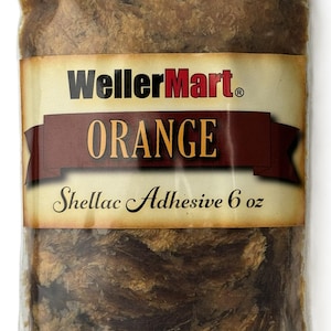 May include: A clear plastic bag filled with orange shellac flakes. The bag has a label that reads "WellerMart" in black text with a red "Mart" and "ORANGE" in black text. The label also says "Shellac Adhesive 6 oz" in black text.