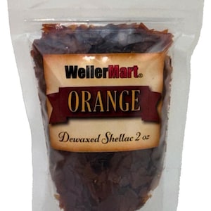 May include: A clear plastic bag filled with orange-brown flakes of dewaxed shellac. The bag has a label that reads "WellerMart" and "ORANGE Dewaxed Shellac 2 oz".