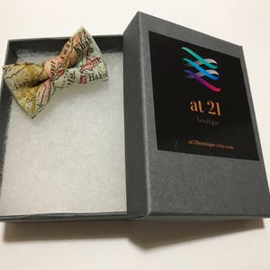 May include: A gray gift box with a white and light blue bow tie. The bow tie is made of fabric with a map print. The box has a logo that says "at 21 boutique" and a website address.