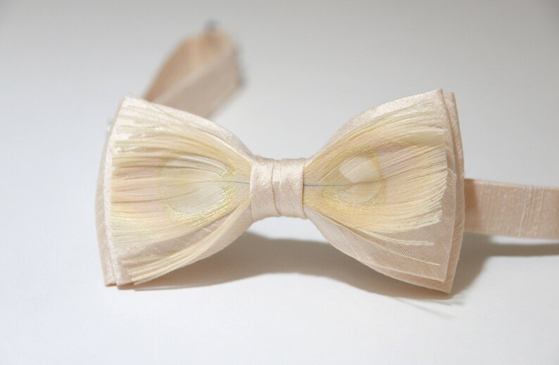 May include: A cream-colored bow tie made of feathers with a subtle shimmer. The bow tie is tied and ready to wear.