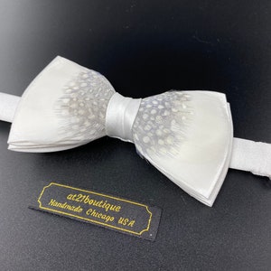 May include: White bow tie with a gray and white feather pattern. The bow tie is handmade in Chicago, USA. The label reads "at21boutique Handmade Chicago USA".