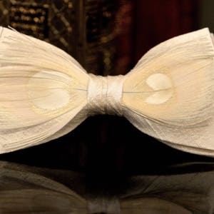 May include: A white bow tie made of feathers. The bow tie has a subtle pattern of light brown spots.