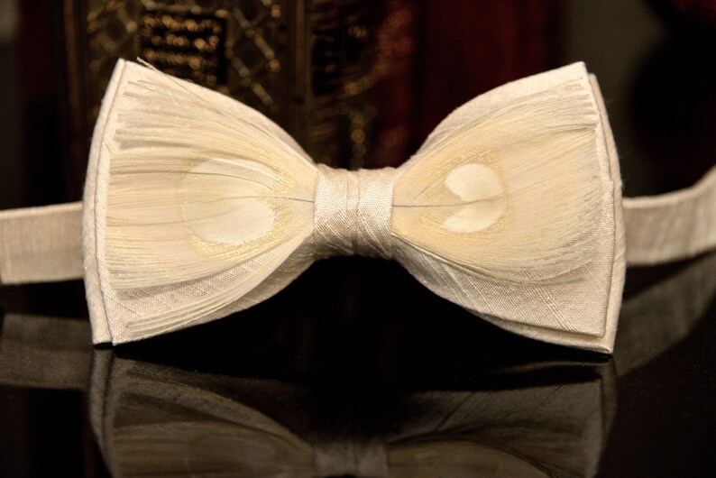 May include: A white bow tie made of feathers. The bow tie has a subtle pattern of light brown spots.