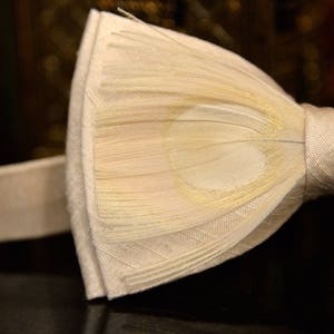 May include: A white bow tie made of feathers with a satin band. The bow tie is tied and ready to wear.