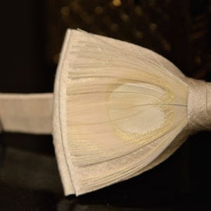 May include: A white bow tie with a textured, layered design. The bow tie has a subtle gold sheen and is tied with a white satin ribbon.