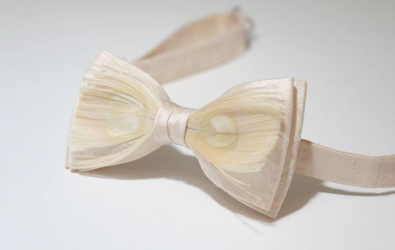 May include: A cream-colored bow tie made of layered fabric with a subtle shimmer. The bow tie has a simple design and is perfect for a formal occasion.