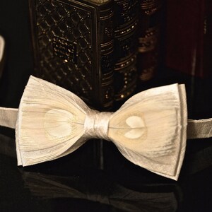 May include: A white bow tie made of layered fabric with a subtle sheen. The bow tie is tied and ready to wear.