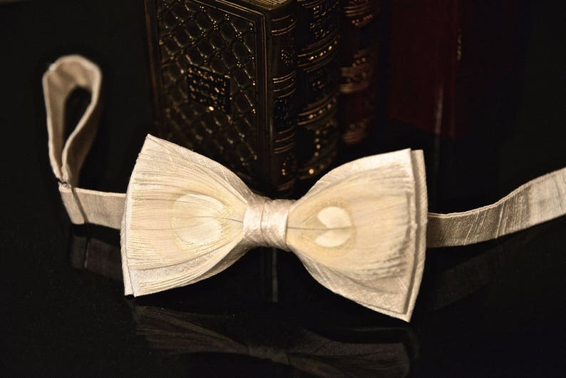 May include: A white bow tie made of layered fabric with a subtle sheen. The bow tie is tied and ready to wear.