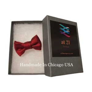 May include: A small, dark red bow tie in a gray gift box with white tissue paper. The box has a label with the text "at 21 boutique" and a website address. The text "Handmade in Chicago USA" is printed on the box.