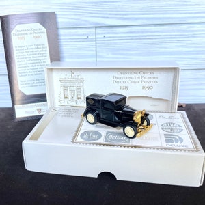 May include: A black vintage car model with gold accents and cream-colored wheels, displayed on a white box with the text "Deluxe Check Printers 1915-1990". The box also features the company's logo and a brochure.