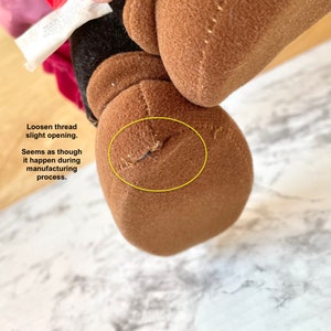 May include: Close-up of a brown plush toy with a loose thread near the seam. The thread appears to have come loose during the manufacturing process.