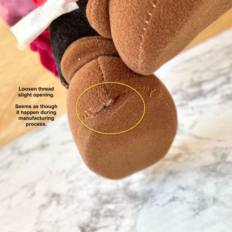 May include: Close-up of a brown plush toy with a loose thread near the seam. The thread appears to have come loose during the manufacturing process.