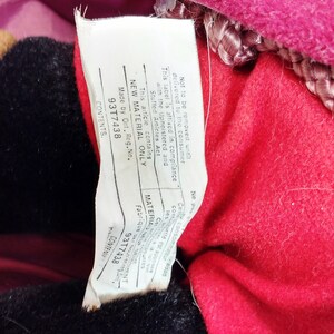 May include: White tag with black text attached to a red and black stuffed toy. The tag states "Not to be removed until delivered to the consumer. This label is affixed in compliance with the Upholstered and Stuffed Articles Act. This article contains NEW MATERIAL ONLY. Made by Ont. Reg. No. 93T7438. CONTENTS."