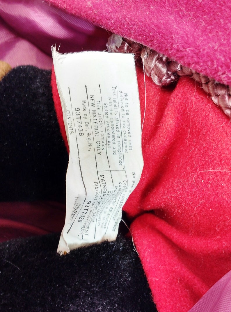 May include: White tag with black text attached to a red and black stuffed toy. The tag states "Not to be removed until delivered to the consumer. This label is affixed in compliance with the Upholstered and Stuffed Articles Act. This article contains NEW MATERIAL ONLY. Made by Ont. Reg. No. 93T7438. CONTENTS."